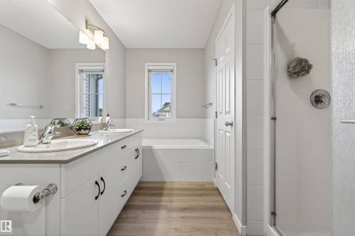 Bathroom featuring a dual-sink vanity with white cabinetry and dark hardware, a built-in bathtub, a walk-in shower with a glass enclosure, wood-finish flooring, and two windows - 22638 99A Avenue, Edmonton, AB 