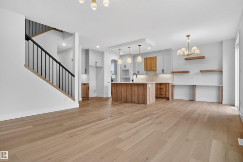 Open-concept living area featuring light wood-finish flooring, a modern kitchen island with pendant lighting, a black iron staircase railing, light-toned cabinetry, and built-in shelving - 15 Redbud Way, St. Albert, AB - Indoor Photo Showing Kitchen