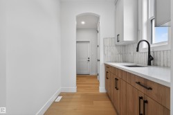 Contemporary wet bar featuring a single basin sink with a matte black faucet, light wood-finish lower cabinetry, white upper cabinetry, and a light-toned countertop - 