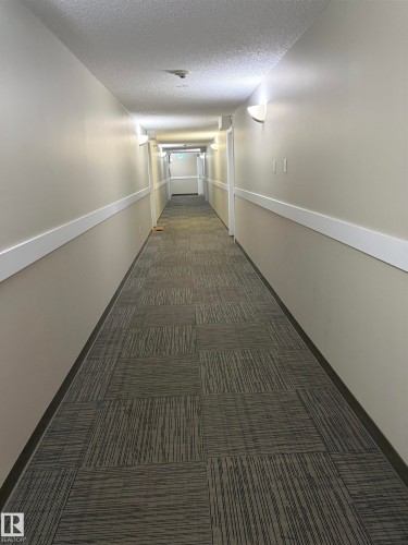 Long corridor with patterned carpet flooring, neutral-toned walls, and wall-mounted sconce lighting - 317 6921 199 Street, Edmonton, AB 