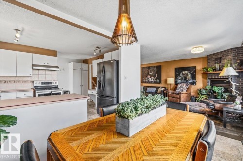 Open-concept living area featuring a wood-finish dining table with a herringbone pattern, a kitchen with white cabinetry and dark appliances, and a living room with a brick fireplace - 135 Homestead Crescent Nw, Edmonton, AB 