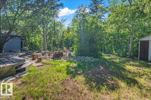 Expansive wooded backyard featuring a raised wood deck, a rock-lined fire pit area with tree stump seating, and a utility shed - 135 Homestead Crescent Nw, Edmonton, AB 