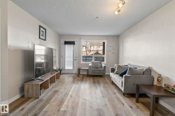 Spacious living area featuring wood-finish flooring, neutral wall tones, a large window, and a white exterior door - 