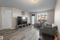 Spacious living area featuring light-toned walls, wood-finish flooring, and ample natural light from a large window - 