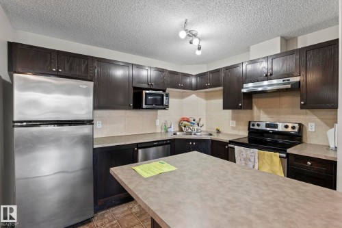 Kitchen featuring dark wood-finish cabinetry, stainless steel appliances, a tile backsplash, and a central island with a solid surface countertop - 2106 9357 Simpson Dr Nw, Edmonton, AB - Indoor Photo Showing Kitchen With Stainless Steel Kitchen With Double Sink With Upgraded Kitchen