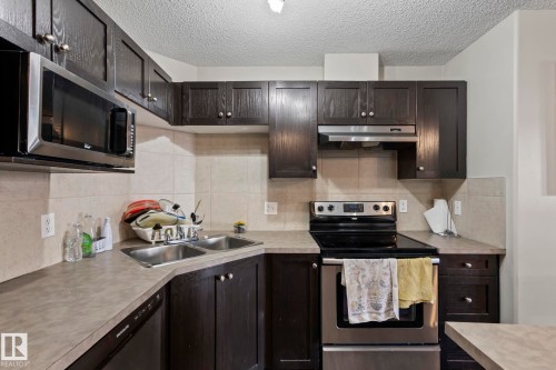 Kitchen featuring dark wood-finish cabinetry, stainless steel appliances, a double basin sink, and neutral tile backsplash - 2106 9357 Simpson Dr Nw, Edmonton, AB - Indoor Photo Showing Kitchen With Stainless Steel Kitchen With Double Sink