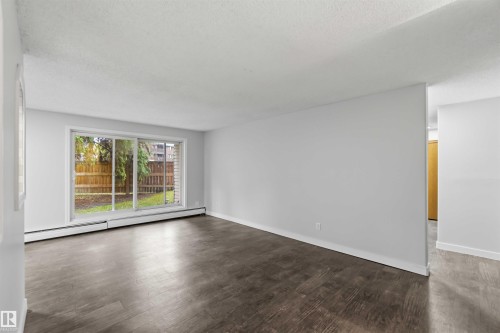 Spacious room featuring wood-finish flooring, a large sliding glass door, white baseboards, and a neutral wall color - 121 5125 Riverbend Road Nw, Edmonton, AB 