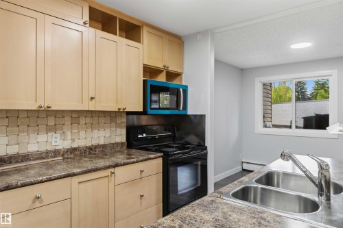 Kitchen featuring light wood-finish cabinetry, patterned tile backsplash, and dark patterned countertops - 121 5125 Riverbend Road Nw, Edmonton, AB 