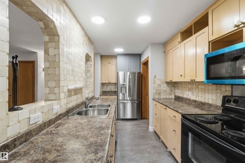 Kitchen featuring stainless steel appliances, dual basin sink, light wood cabinetry, stone-patterned countertops, and a tiled backsplash - 121 5125 Riverbend Road Nw, Edmonton, AB 