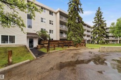 Multi-story building featuring balconies with metal railings, a paved parking area, and surrounding green spaces with mature evergreen trees - 