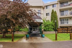 Building exterior featuring a covered entrance, multiple balconies with white railings, and a landscaped frontage with mature trees and shrubs - 