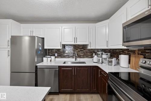 Kitchen featuring stainless steel appliances, white upper cabinetry, rich brown lower cabinetry, a mosaic tile backsplash, and light-toned countertops - 138 16035 132 Street, Edmonton, AB 