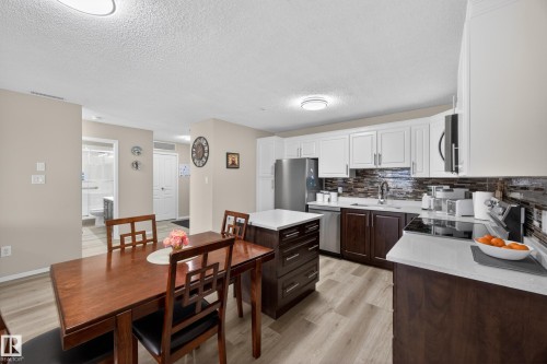 Open-concept kitchen and dining area featuring a central island with countertop seating, stainless steel appliances, two-tone cabinetry, and wood-finish flooring - 138 16035 132 Street, Edmonton, AB 