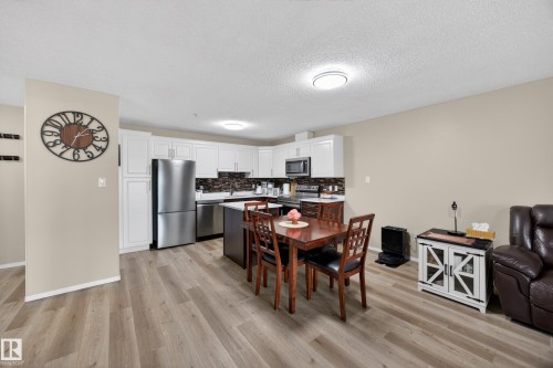 Open-concept kitchen and living area featuring wood-finish flooring, white cabinetry, a dark tile backsplash, stainless steel appliances, and a central island - 138 16035 132 Street, Edmonton, AB 