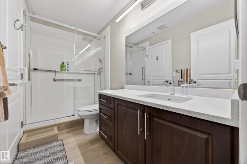 Spacious bathroom featuring a glass-enclosed shower with white subway tile, a dark wood-finish vanity with a light-toned countertop, a rectangular undermount sink, and a large mirror - 138 16035 132 Street, Edmonton, AB 