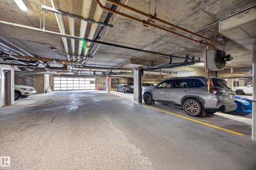 Underground parking garage featuring exposed concrete ceiling, overhead utility piping, and fluorescent lighting - 138 16035 132 Street, Edmonton, AB 