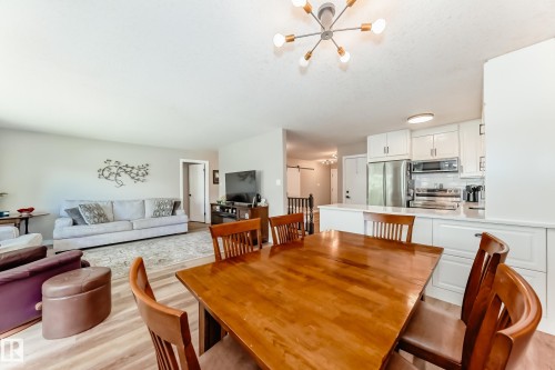 Open-concept living area featuring light wood-finish flooring, a contemporary ceiling light fixture, white kitchen cabinetry with stainless steel appliances, and a breakfast bar with a solid surface countertop - 10822 11 Avenue, Edmonton, AB 