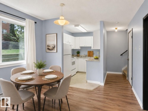 Open-concept living space featuring wood-finish flooring, a kitchen with white cabinetry and appliances, a dining area with a modern pendant light, and a window with exterior green space - 1 13450 Fort Road, Edmonton, AB 