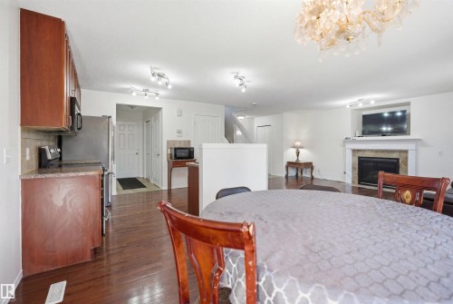 Open-concept living area featuring rich wood-finish flooring, a built-in electric fireplace with stone surround and white mantel, a kitchen with wood cabinetry and dark countertops, and a decorative overhead light fixture - 1560 37B Avenue, Edmonton, AB 