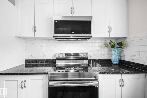 Modern kitchen featuring a stainless steel range with overhead microwave, white cabinetry with silver hardware, black speckled countertops, and a white subway tile backsplash - 4806 117 Ave, Edmonton, AB 