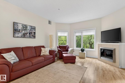 Living area with wood-finish flooring, two large windows, a corner fireplace with a tiled surround, and neutral wall paint - 208 5 Gate Avenue, St. Albert, AB 