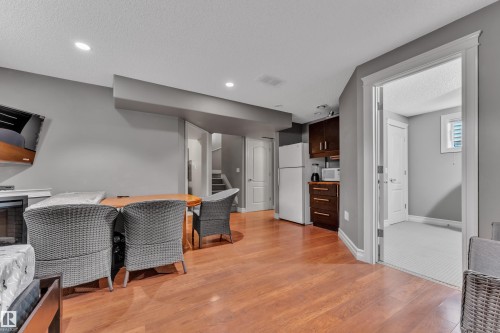 Wood-finish flooring throughout a versatile living area with a built-in kitchen featuring dark wood cabinetry and a white refrigerator - 6116 11 Avenue, Edmonton, AB 