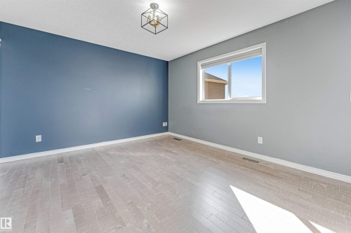 Spacious room featuring light wood-finish flooring, a contemporary ceiling light fixture, and a large window with white trim - 5928 12 Avenue, Edmonton, AB 