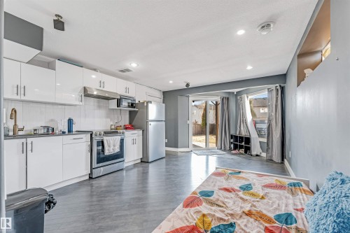 Open-concept living space featuring a full kitchen with white cabinetry, stainless steel appliances, and dark countertops - 5928 12 Avenue, Edmonton, AB 