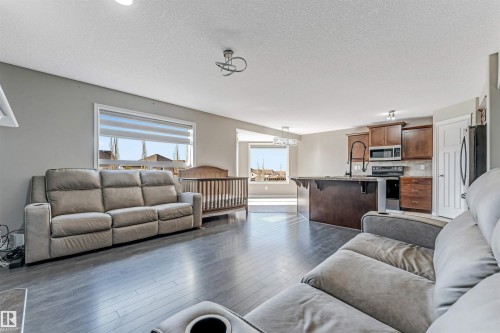 Open-concept living space featuring wood-finish flooring, a contemporary ceiling light fixture, and a kitchen with dark cabinetry, stainless steel appliances, and a central island with a stone countertop - 5928 12 Avenue, Edmonton, AB 