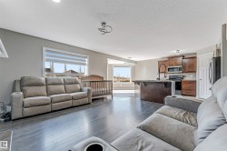Open-concept living space featuring wood-finish flooring, a contemporary ceiling light fixture, and a kitchen with dark cabinetry, stainless steel appliances, and a central island with a stone countertop - 