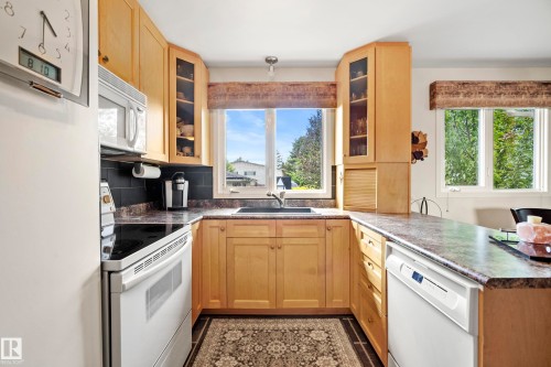U-shaped kitchen featuring light wood cabinetry, dark countertops, a black tile backsplash, and white appliances including a range, microwave, and dishwasher - 3208 70 Street, Edmonton, AB 