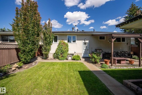 Expansive backyard featuring a green lawn, concrete pathway, mature trees, and a covered patio with a wooden pergola - 3208 70 Street, Edmonton, AB 