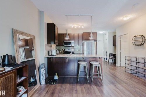 Open-concept kitchen featuring dark wood cabinetry, stainless steel appliances, a light-toned tile backsplash, and a breakfast bar with pendant lighting - 217 508 Griesbach Parade, Edmonton, AB 