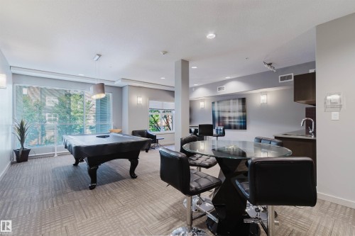 Spacious recreation room featuring patterned carpet flooring, a large window offering natural illumination, recessed lighting, and a built-in wet bar with dark cabinetry and a stainless steel sink - 217 508 Griesbach Parade, Edmonton, AB 