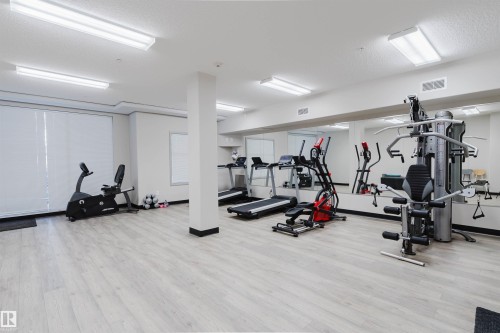Spacious fitness center featuring light wood-finish flooring, full-wall mirrors, abundant overhead lighting, and white window blinds - 217 508 Griesbach Parade, Edmonton, AB 