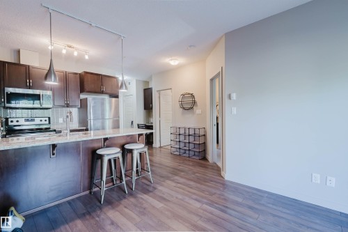 Kitchen featuring dark wood cabinetry, stainless steel appliances, a tile backsplash, and a breakfast bar with pendant lighting - 217 508 Griesbach Parade, Edmonton, AB 