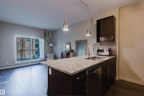 Kitchen island featuring granite countertops, undermount sink with gooseneck faucet, dark wood-finish cabinetry, and stainless steel dishwasher - 217 508 Griesbach Parade, Edmonton, AB 