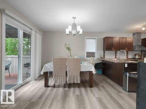 Open-concept living space featuring wood-finish flooring, a sliding glass door to an outdoor deck, a modern chandelier, and a kitchen area with brown cabinetry, a stainless steel range hood, and a kitchen sink with a gooseneck faucet - 3756 8 Street, Edmonton, AB 