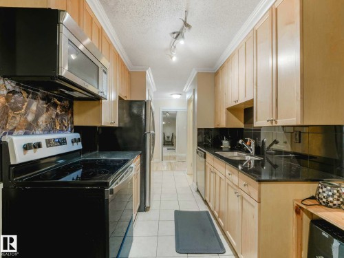 Galley kitchen featuring light wood-finish cabinetry, black countertops, stainless steel microwave, black electric range, and black refrigerator - 403 10165 113 Street, Edmonton, AB 