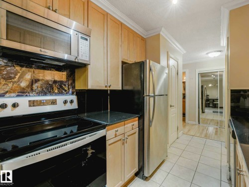 Kitchen featuring light wood-finish cabinetry, stainless steel appliances, black countertops, and a tiled floor - 403 10165 113 Street, Edmonton, AB 