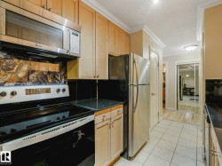Kitchen featuring light wood-finish cabinetry, stainless steel appliances, black countertops, and a tiled floor - 