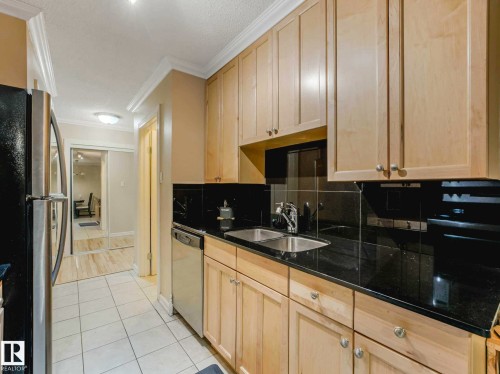 Kitchen featuring natural wood cabinetry, dark countertops, a stainless steel double-basin sink, and a black tile backsplash - 403 10165 113 Street, Edmonton, AB 