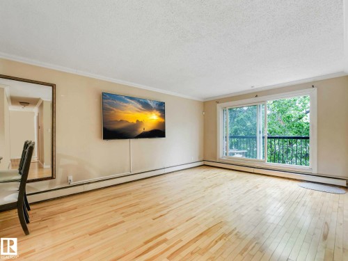 Expansive room featuring light wood-finish flooring, a full-wall mirror, and a sliding glass door opening to a balcony with black railings - 403 10165 113 Street, Edmonton, AB 