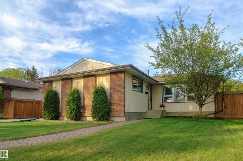 Exterior featuring a brick facade with vertical siding accents, a paved walkway, established lawn, mature tree, and a wooden fence - 58 Lodgepole Crescent, St. Albert, AB 