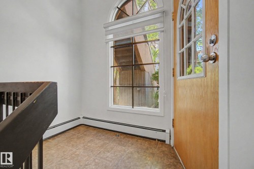 Entryway featuring a wood-panel door with arched glass inserts, an adjacent arched window, tile flooring, white baseboards, and a dark wood staircase railing - 11150 83 Avenue, Edmonton, AB 