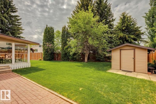 Spacious backyard featuring a manicured lawn, a red paver patio, a sunroom with white railing, a storage shed, and mature perimeter trees - 15804 80 Street, Edmonton, AB 