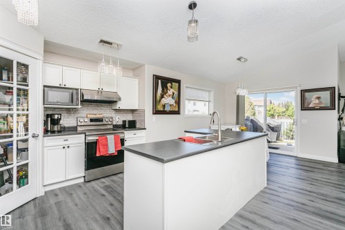 Kitchen featuring a central island with an integrated double sink, white cabinetry with dark hardware, stainless steel appliances, a tiled backsplash, and wood-finish flooring - 4228 162 Avenue, Edmonton, AB 