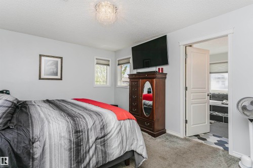 Plush carpeted room featuring a contemporary chandelier, two windows with roller shades, and an open doorway revealing a bathroom with hexagon floor tiles and a built-in tub - 4228 162 Avenue, Edmonton, AB 