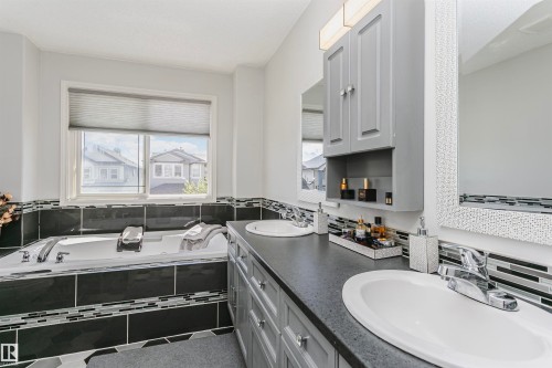 Bathroom featuring a built-in tub with black and mosaic tile surround, dual vanity with undermount sinks and dark countertops, shaker-style cabinetry, and a window with privacy blinds - 4228 162 Avenue, Edmonton, AB 