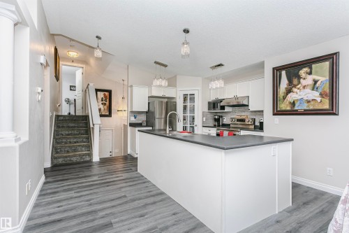 Open-concept kitchen featuring an island with a sink, stainless steel appliances, white cabinetry, wood-finish flooring, and pendant lighting - 4228 162 Avenue, Edmonton, AB 